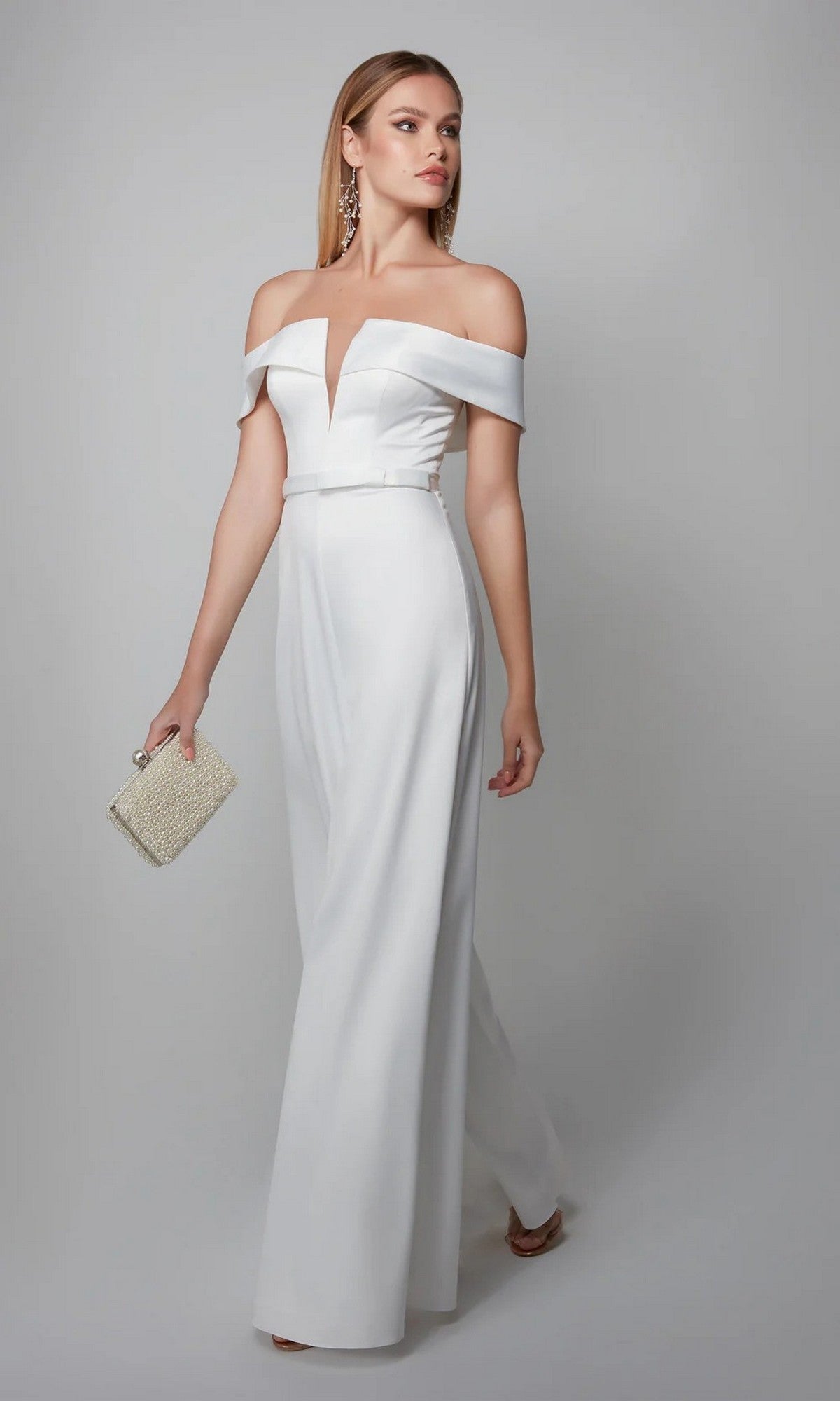 A woman in the Alyce 70012 long formal jumpsuit in diamond white holds a small beaded clutch, standing against a plain light gray background.
